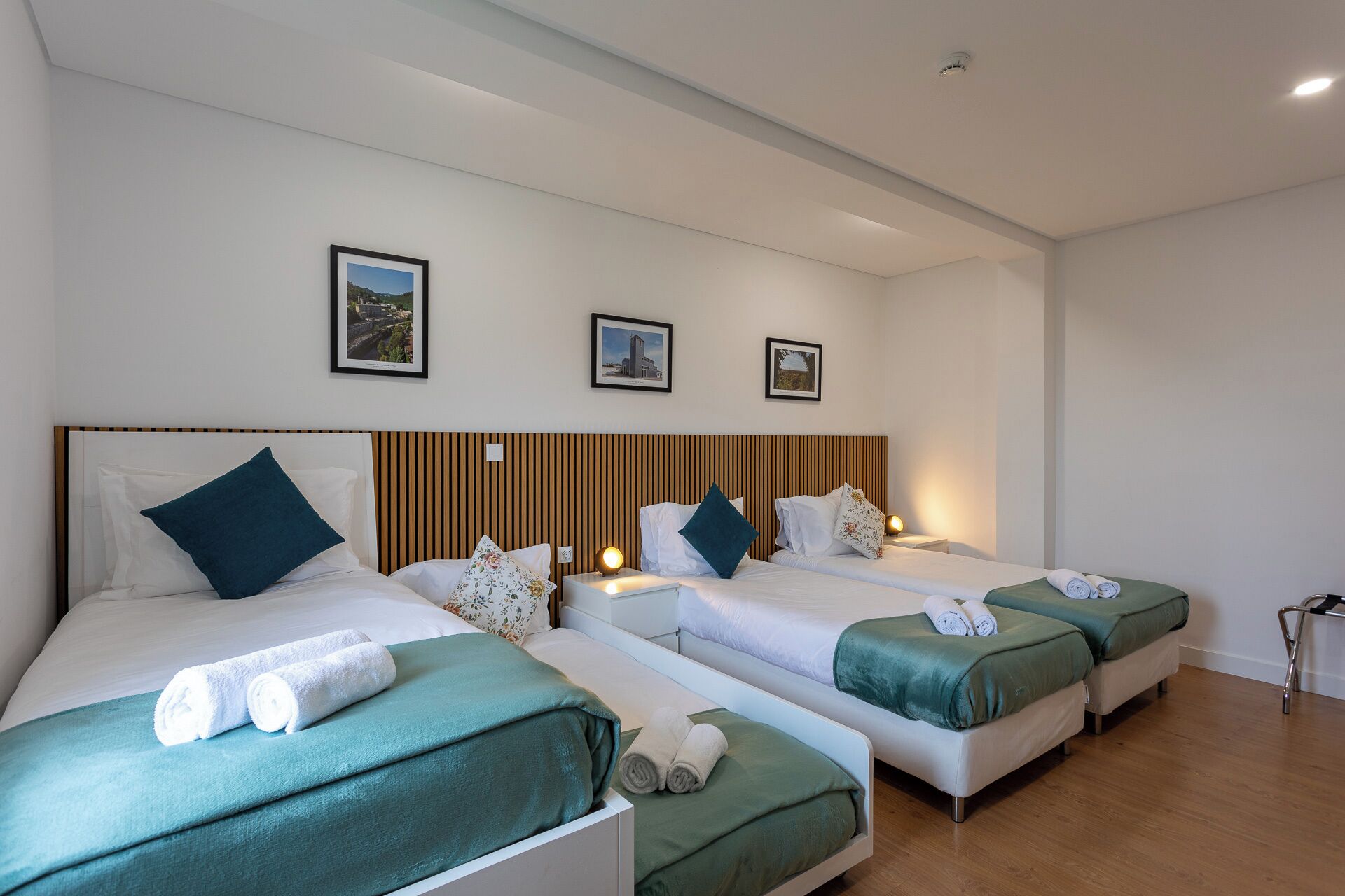 Een hotelkamer met drie eenpersoonsbedden, elk met een groene sprei en witte kussens. Boven de bedden hangen afbeeldingen in lijsten.