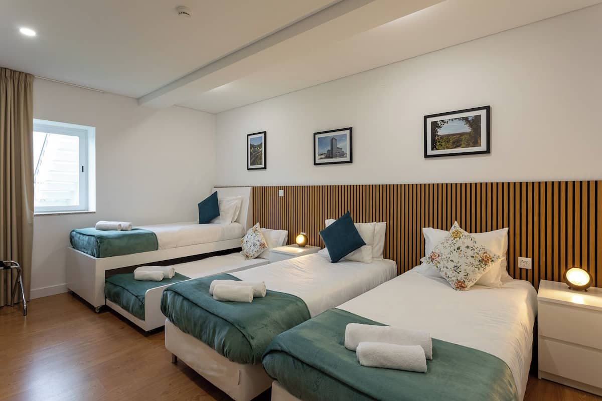 A hotel room with three beds, each with green bedding and white pillows. There are framed pictures on the wall, a window with curtains, and a wooden headboard.