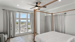 Ein Schlafzimmer mit Himmelbett, großen Fenstern mit Vorhängen, Deckenventilator und Blick ins Freie.