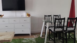 Una habitación con una televisión de pantalla plana montada en la pared, un aparador blanco con cajones, una mesa de comedor con superficie de vidrio y cuatro sillas, y un taburete pequeño.