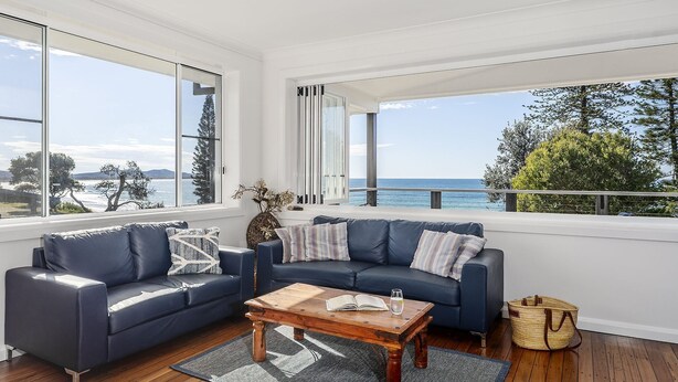 A modern living room with a blue sofa, a wooden coffee table, and large windows offering a view of the sea. - Aqua Vista - Beach Front Location (Brooms Head)