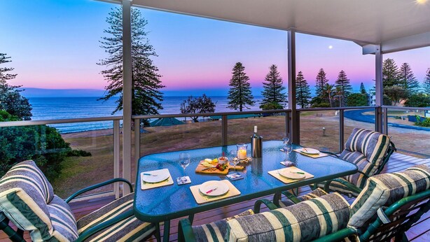 A dining area with a table set for two, overlooking a beach and ocean at sunset. - Casa della vista - Beach Front Location (Brooms Head)
