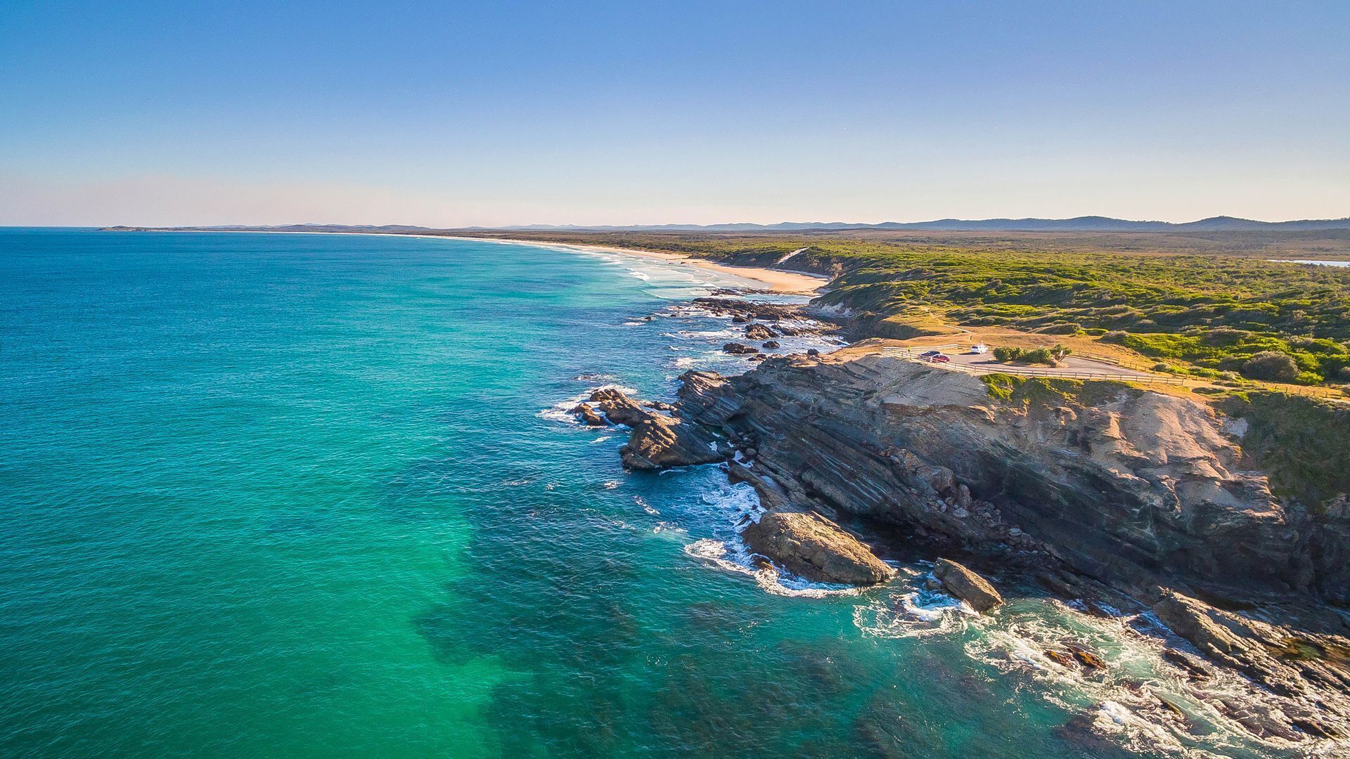 A coastal landscape with clear turquoise waters, rocky cliffs, and a distant view of a beach with sparse vegetation.