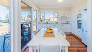 A modern kitchen with white cabinets, a dining table with a fruit bowl, and a view of the outdoors through large windows. - Casa della vista - Beach Front Location (Brooms Head)
