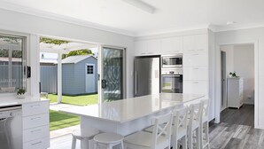 A modern kitchen with white cabinetry, a large island with bar stools, and a view of the backyard. - A Little Beach Haven - In the heart of Brooms Head (Brooms Head)