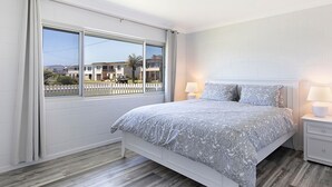 A bedroom with a large window, a bed with patterned bedding, two bedside lamps, and a white nightstand. - A Little Beach Haven - In the heart of Brooms Head (Brooms Head)