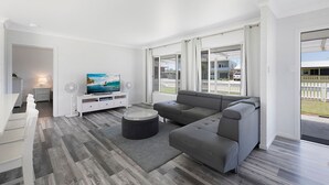 A modern living room with a grey sectional sofa, a flat-screen TV, and two standing fans. - A Little Beach Haven - In the heart of Brooms Head (Brooms Head)