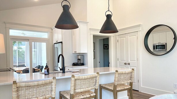 A modern kitchen with white cabinetry, a central island with bar stools, and pendant lighting. - NEW!—IBIS COTTAGE—Beach side retreat. Prime location with Bikes and Beach gear! (Oak Island)