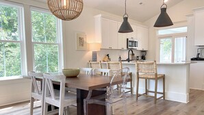 A modern kitchen with white cabinets, a wooden dining table, and clear chairs. - NEW!—IBIS COTTAGE—Beach side retreat. Prime location with Bikes and Beach gear! (Oak Island)