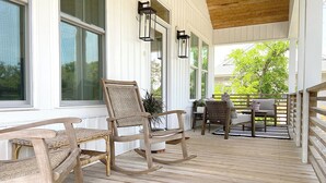 A wooden deck with rocking chairs and a table, surrounded by white walls and large windows. - NEW!—IBIS COTTAGE—Beach side retreat. Prime location with Bikes and Beach gear! (Oak Island)