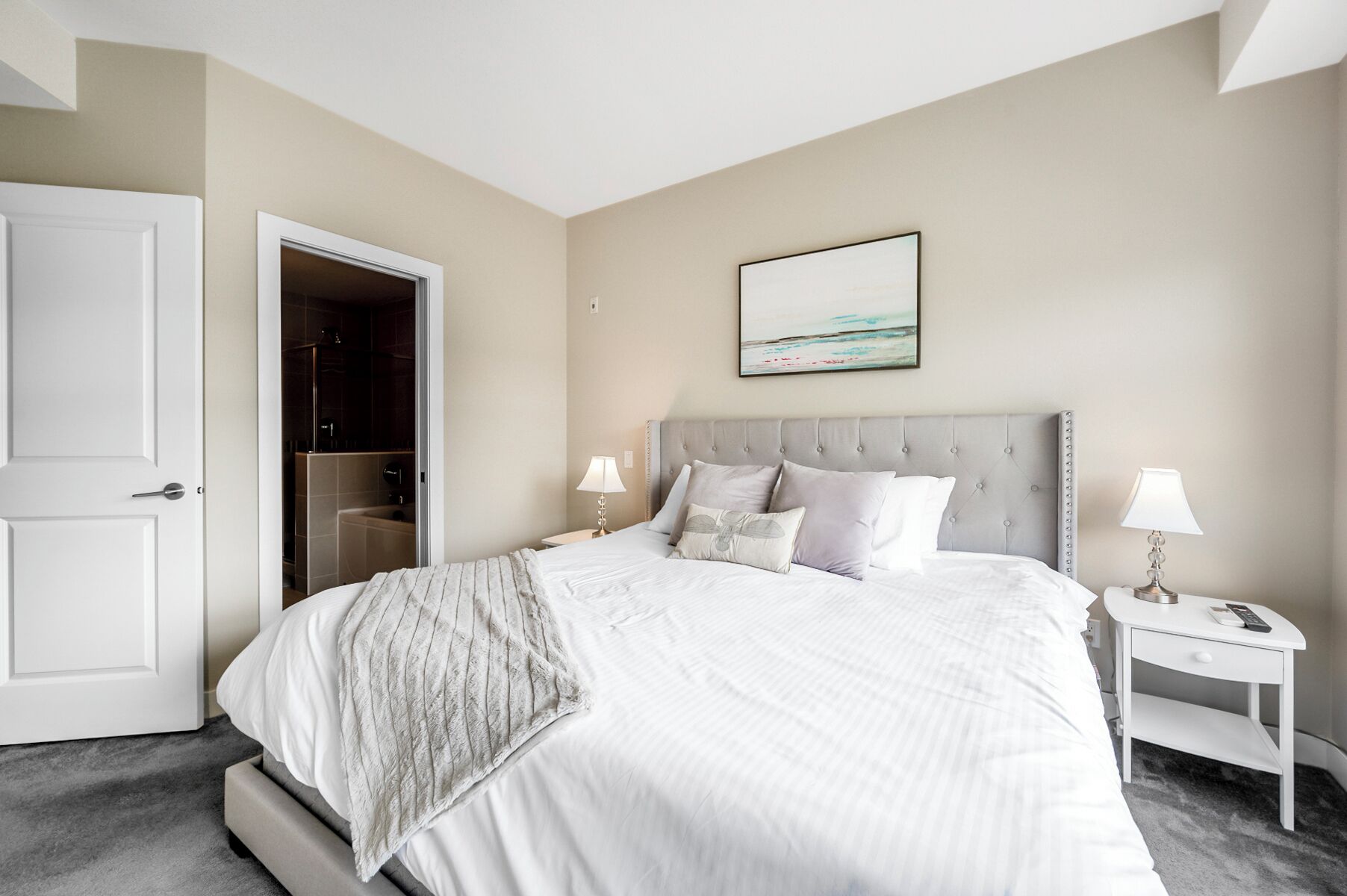 A hotel room with a bed, bedside tables, a nightstand, a painting, and a door leading to a bathroom.