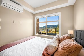 A bedroom with a large bed, a window with a view of the countryside, an air conditioning unit, and a bedside lamp. - Top Floor Condo at Amenity Rich Resort (West Kelowna)