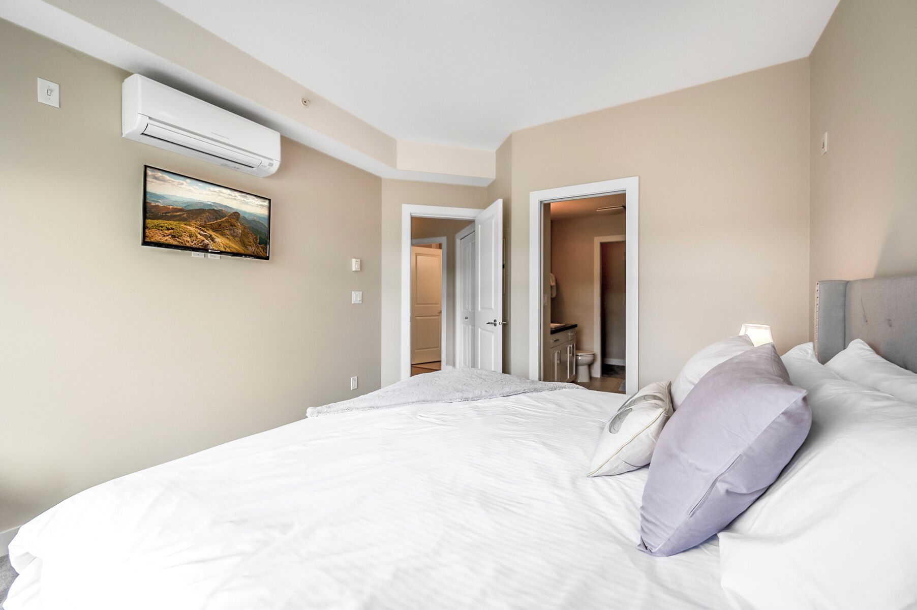 A hotel room with a large bed, a mounted air conditioner, a painting on the wall, and a bathroom visible through an open door.