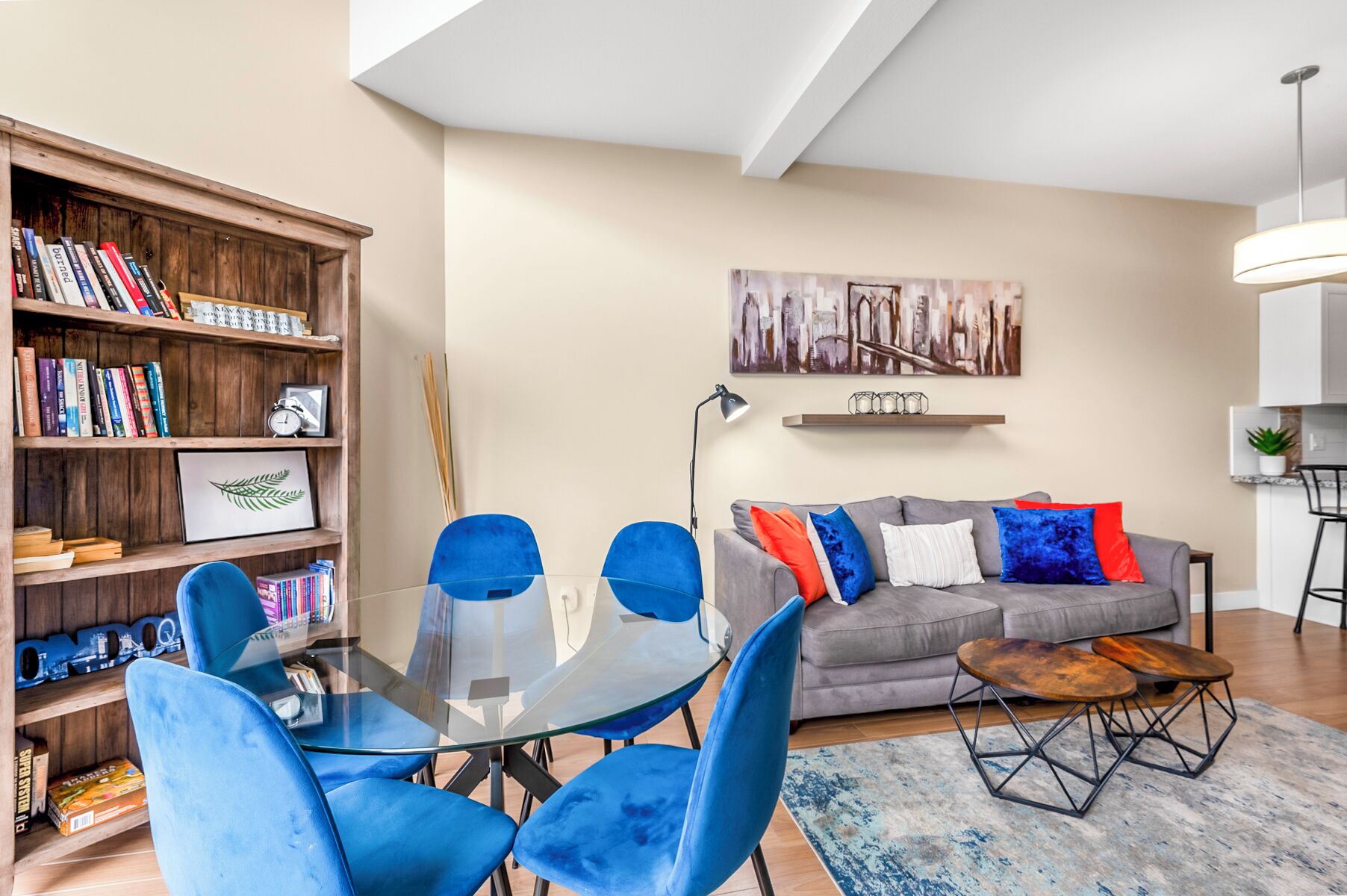 Ein modernes Wohnzimmer mit einem Glastisch, blauen Stühlen, einem grauen Sofa mit bunten Kissen, einem Bücherregal aus Holz und einem Stadtbild an der Wand.