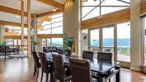 A spacious dining area with a large table and chairs, large windows offering a view of the outdoors, and a staircase leading to an upper level. - Top Floor Condo at Amenity Rich Resort (West Kelowna)