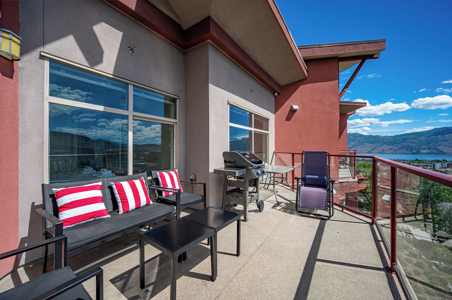 An outdoor balcony with a sofa, table, grill, and a view of a lake and mountains.