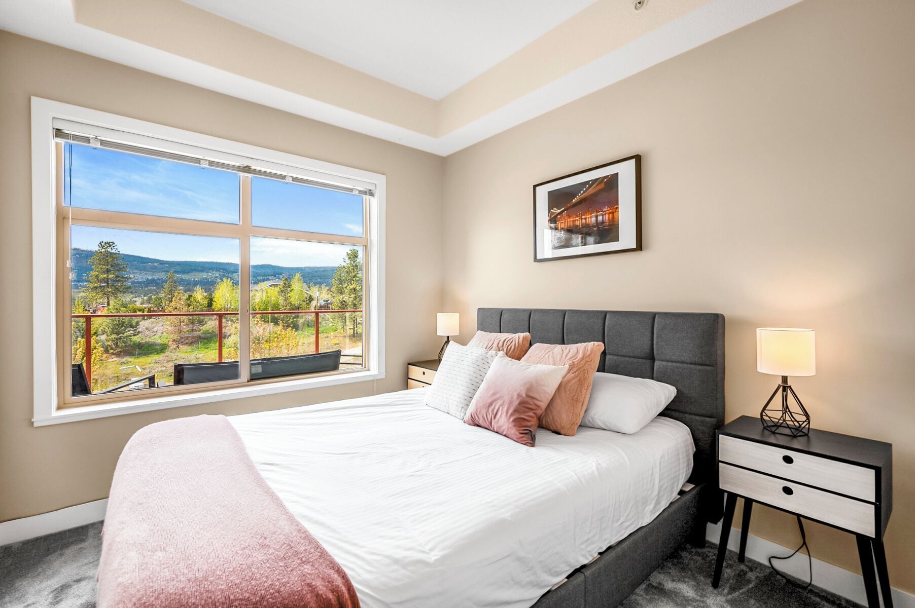 A bedroom with a large window offering a scenic view, a bed with multiple pillows, a bedside table with a lamp, and a framed picture on the wall.