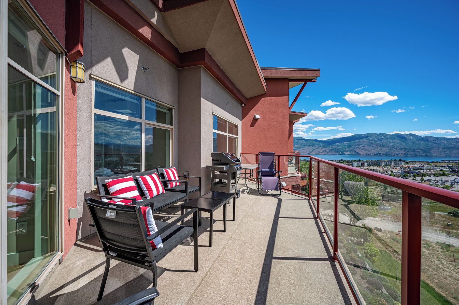 A balcony with a sofa and chairs, a grill, and a view of mountains and a lake.