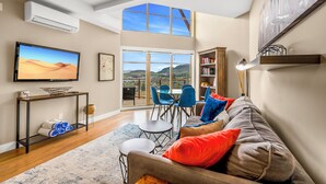 A spacious living area with a large window offering a view of the countryside, a television, a bookshelf, and a cozy seating arrangement. - Top Floor Condo at Amenity Rich Resort (West Kelowna)