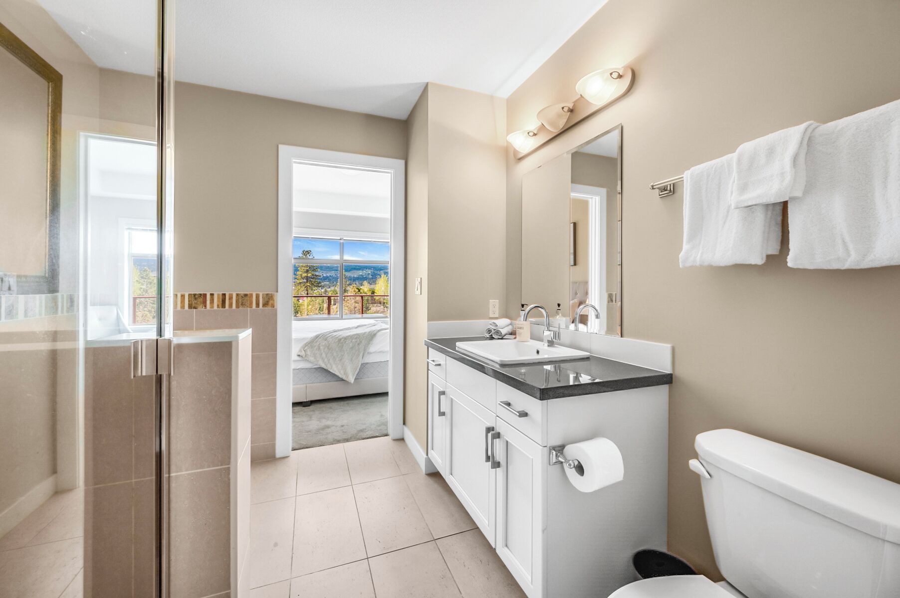 Une salle de bain moderne équipée d’une douche, de toilettes et d’un lavabo. Elle comprend un grand miroir, un porte-serviettes et offre une vue sur un balcon donnant sur un paysage pittoresque.