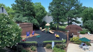 A playground with slides and a brick playhouse, surrounded by trees and a patio area with outdoor seating.