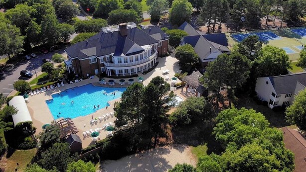 An aerial view of a residential area with a large swimming pool surrounded by lounge chairs and umbrellas, a tennis court, and a large house with a curved roof.