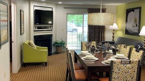 A dining area with a table set for four, chairs with patterned upholstery, a television mounted above the fireplace, and a view of the outside through a glass door.