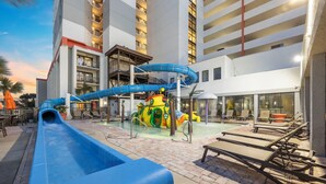 Un espace de parc aquatique comprenant un toboggan bleu, une structure de jeux jaune et verte, et des chaises longues.