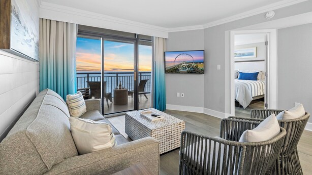 Une chambre d’hôtel moderne avec un balcon, une grande fenêtre donnant sur le coucher de soleil, un lit avec une couvre-lit à rayures et un téléviseur fixé au mur.