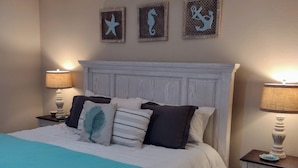 Ein ordentlich gemachtes Bett mit einer türkis farbenen Tagesdecke, zwei Nachttische mit Lampen und drei Wanddekorationen.