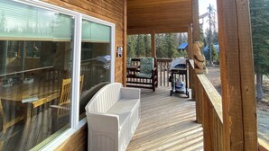 Una terraza de madera con un banco blanco, un comedor con mesa y sillas, y vistas al bosque circundante.