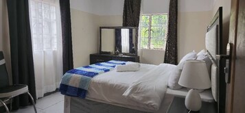 Una habitación de hotel con cama, mesita de noche, espejo, ventana con cortinas y una silla.