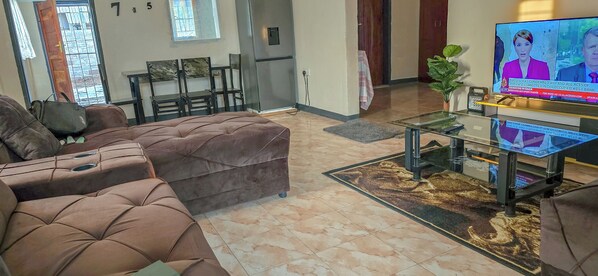 A living room with a brown sofa, a glass coffee table, and a TV displaying a news broadcast. - Eagle Weavers Nest (Livingstone)