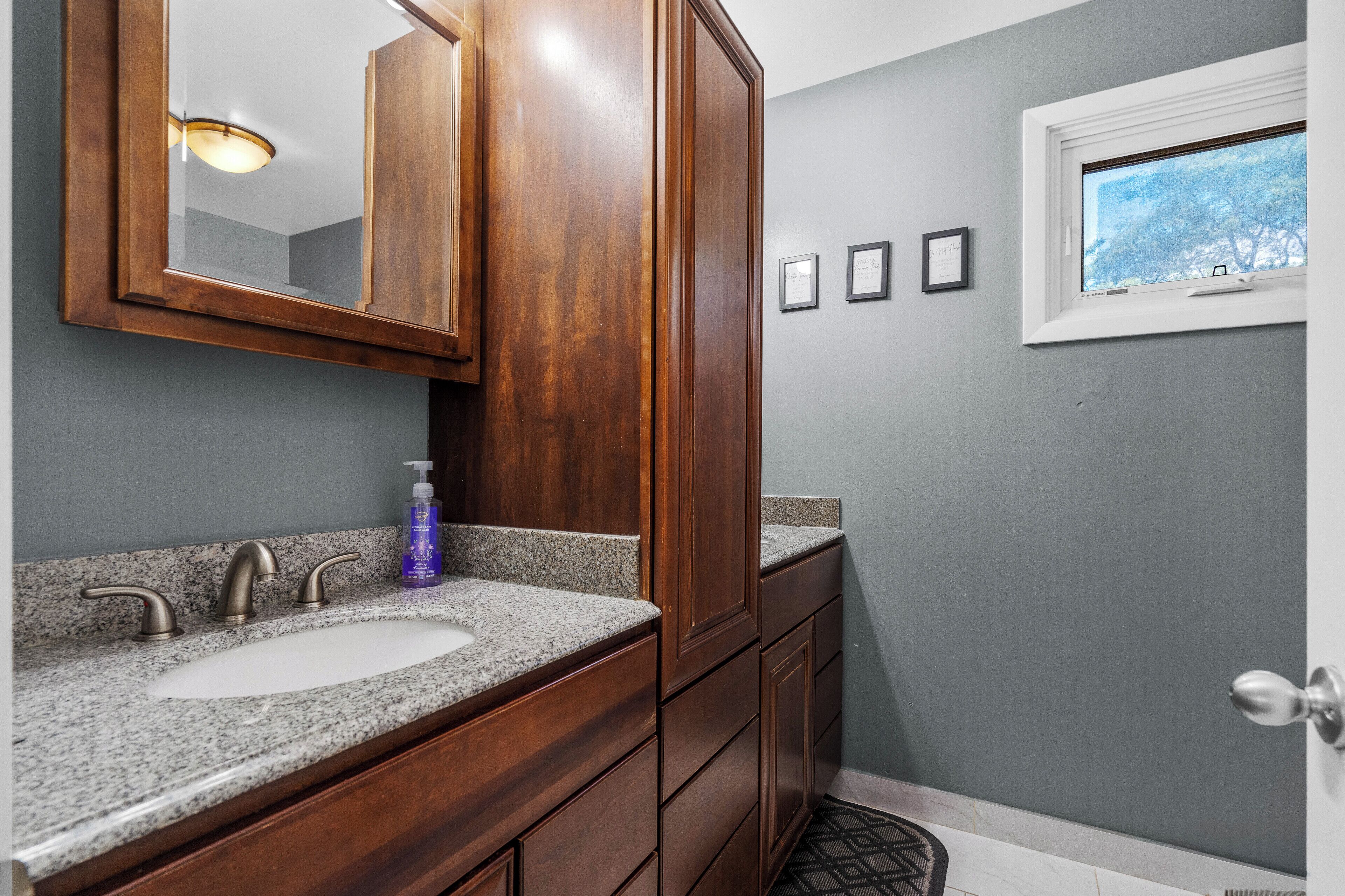 A bathroom with a wooden vanity, a large mirror, a sink, and a window with a view of trees.