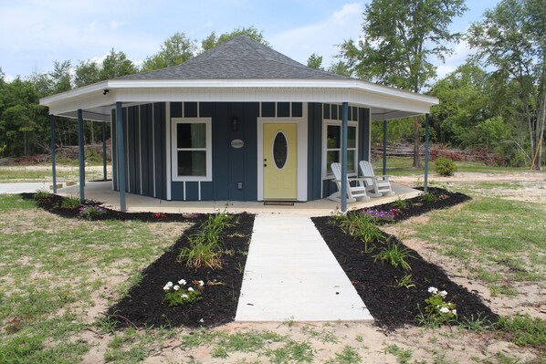 A small, single-story building with a gabled roof, a central entrance door, and a covered porch area with two white chairs. - Revelation House (Eastman)
