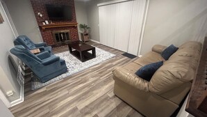 A living room with a brick fireplace, a blue armchair, a brown leather sofa, and a wooden coffee table.