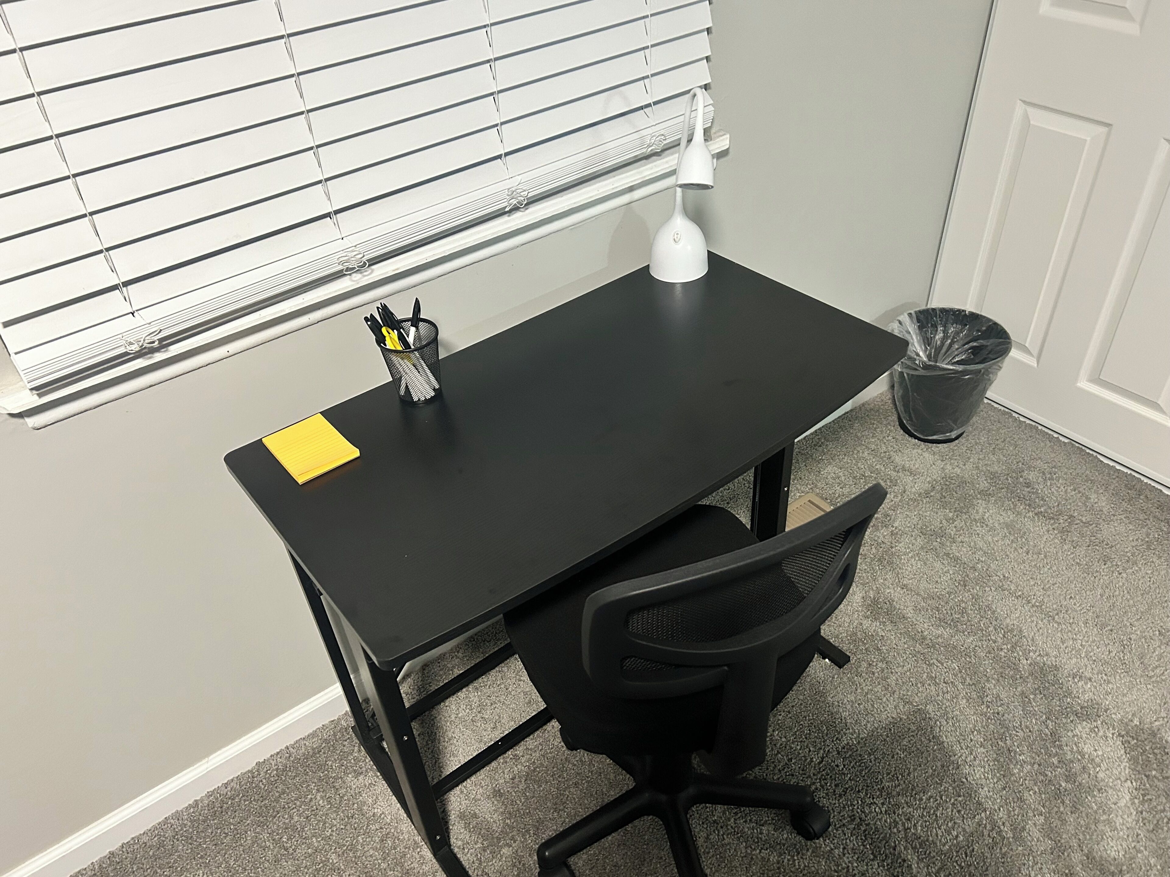 A small office space with a black desk, a chair, a white desk lamp, a trash can, and a window with blinds.