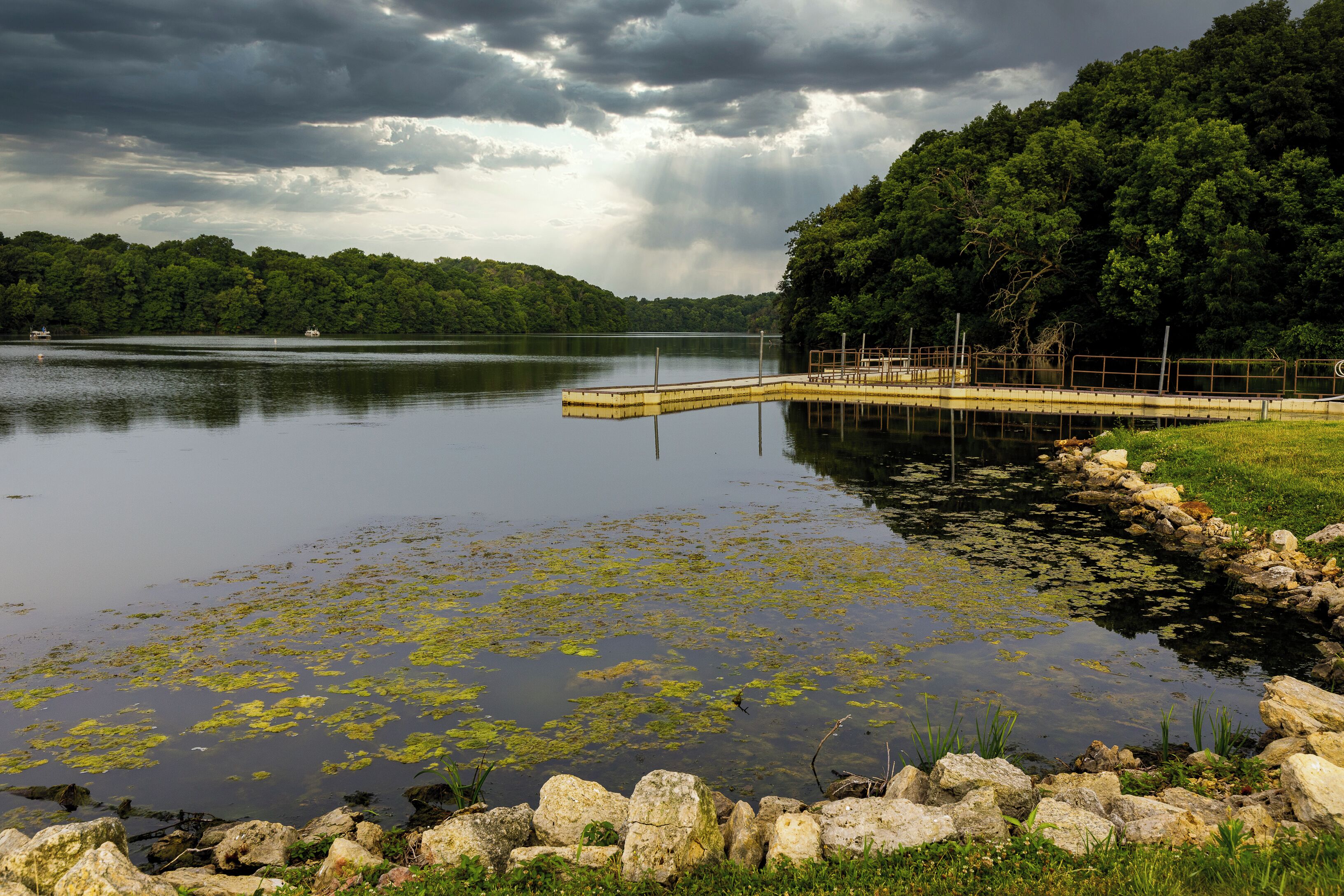 A calm lake with a dock extending into the water, surrounded by lush greenery and a cloudy sky.