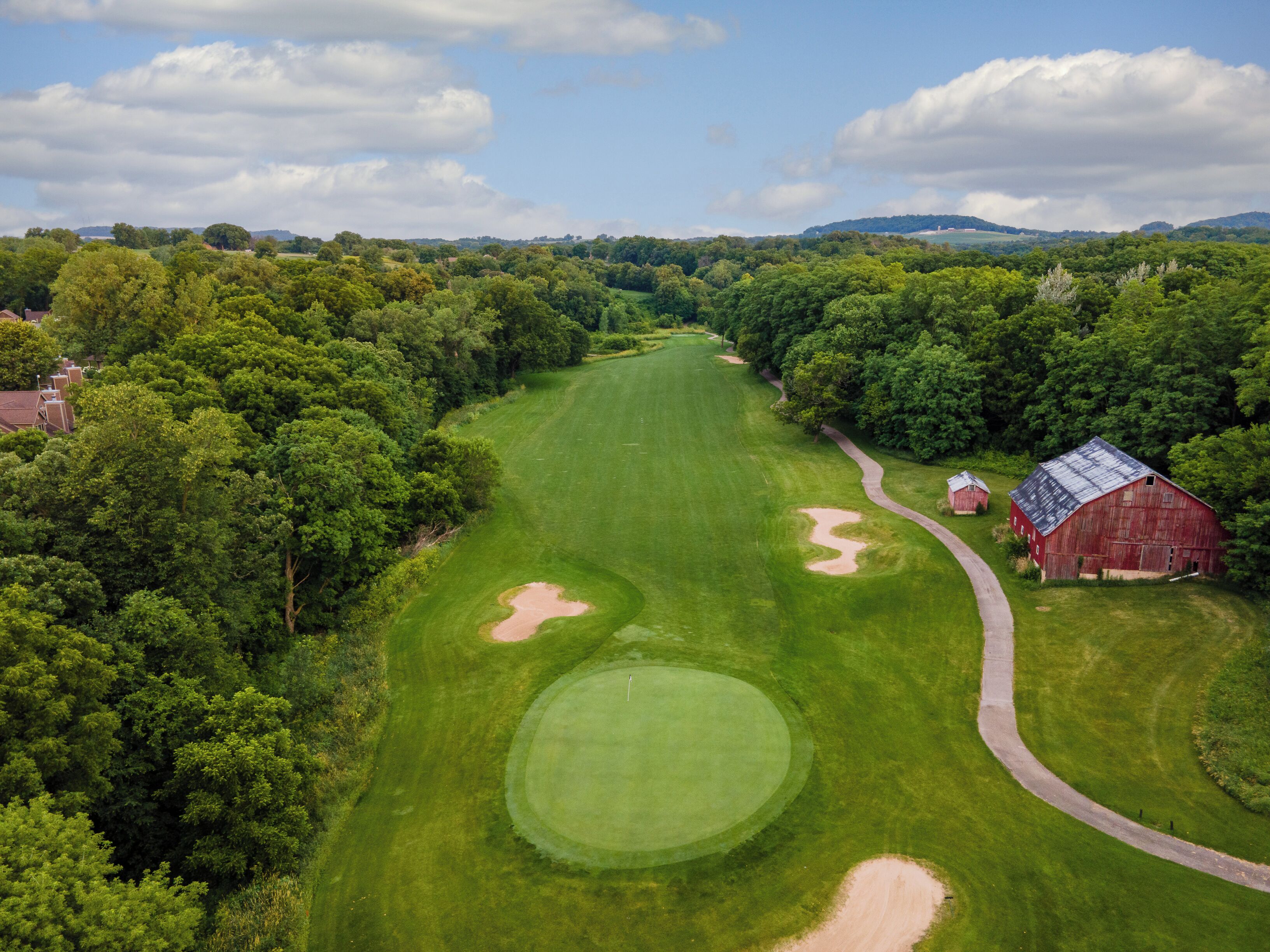 Luftaufnahme eines Golfplatzes mit einer roten Scheune, umgeben von dichten Bäumen und blauem Himmel.