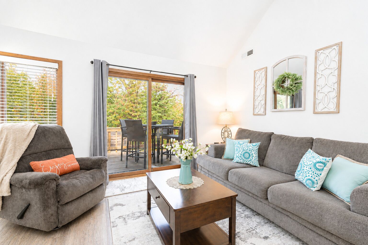 Un salon avec un canapé gris, un fauteuil assorti, une table basse en bois et, à travers une porte vitrée, une vue sur un espace patio extérieur.
