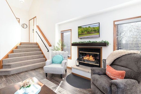 A living room with a fireplace, a television mounted above it, and a staircase leading to another level. - Spacious, King Beds, Pool steps away, Near Golf, Spa (Galena)