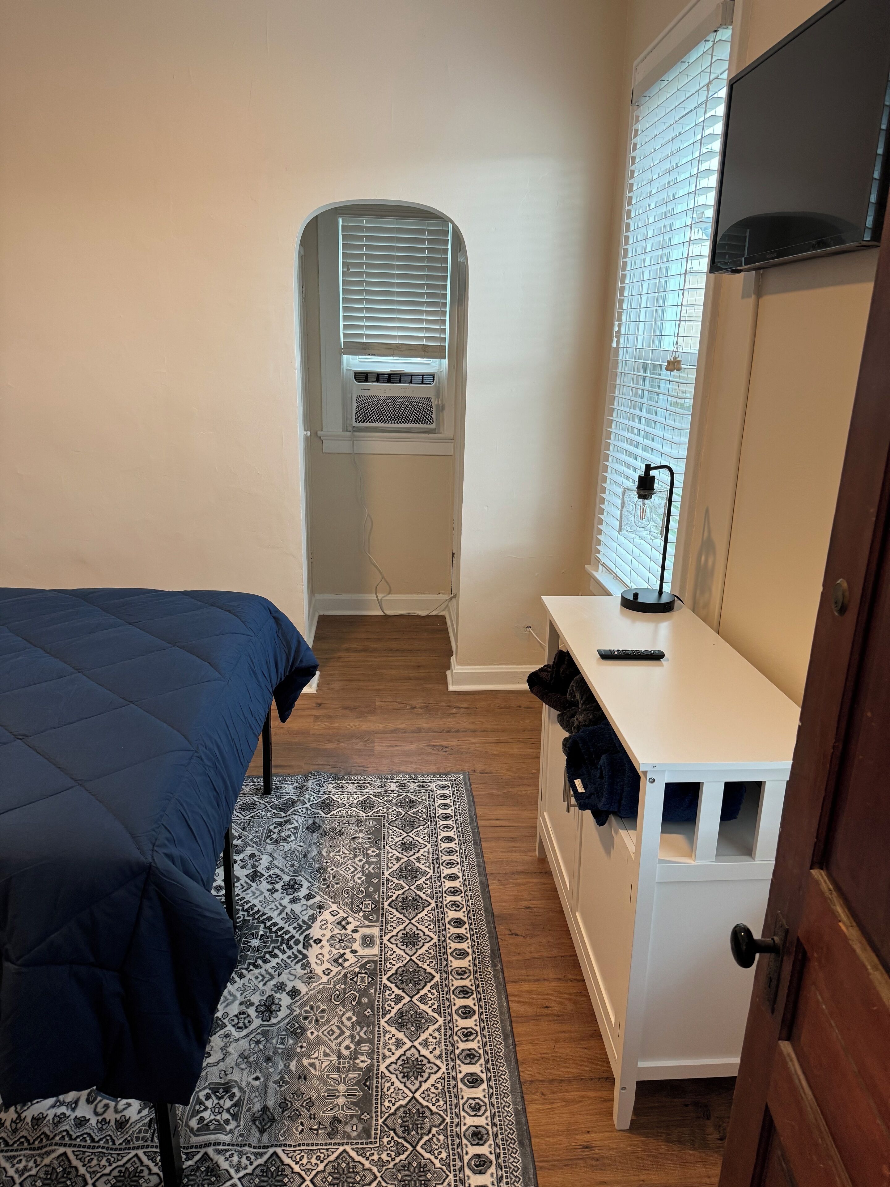 A bedroom with a bed, a desk, a chair, a window with blinds, and an air conditioning unit.