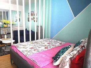 Un dormitorio con cama, escritorio y silla. Hay una ventana con cortinas, una estantería con objetos y una pared con diseño geométrico.