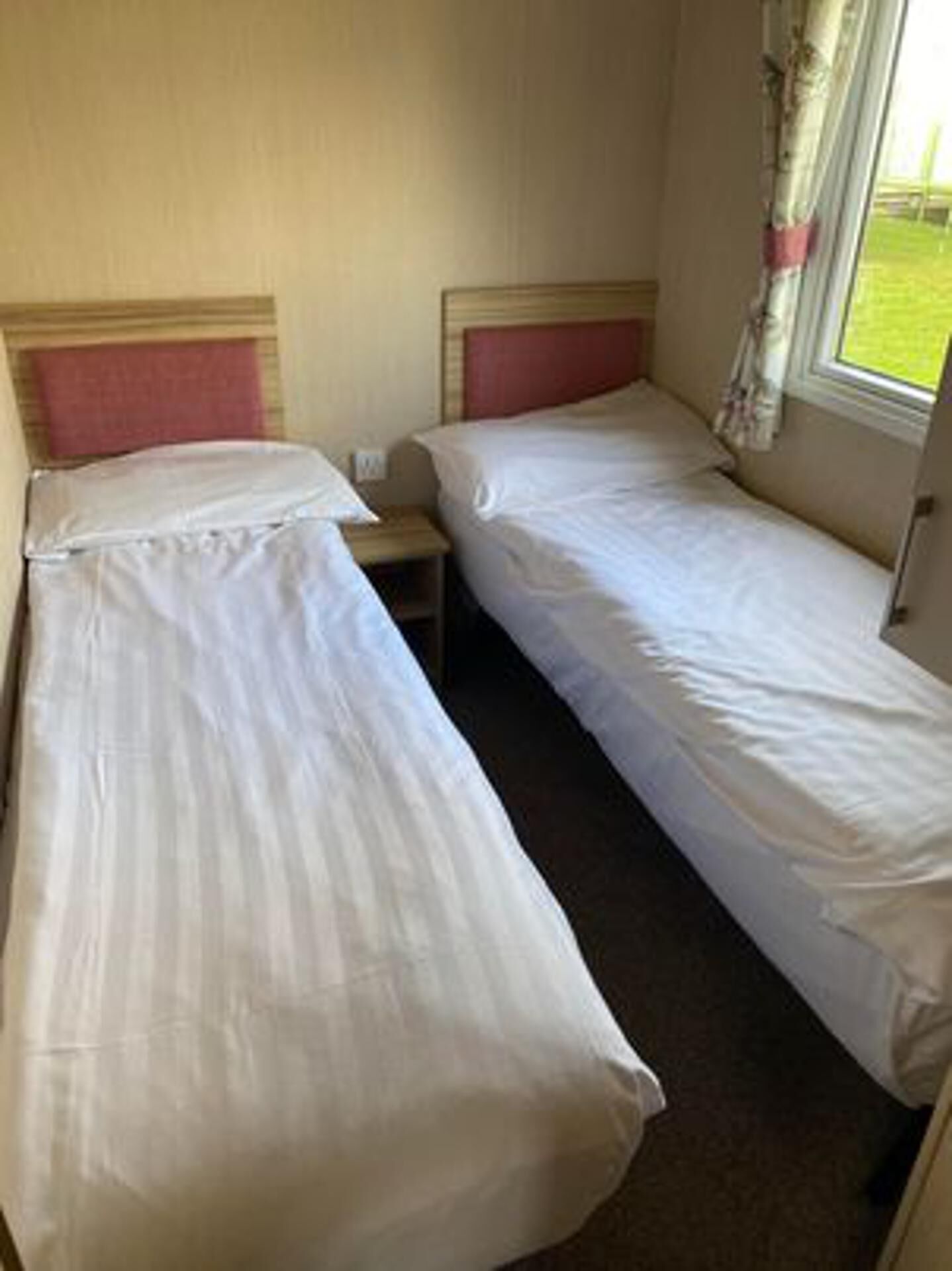 Une petite chambre d’hôtel composée d’une seule pièce, avec deux lits simples, une table de chevet et une fenêtre avec des rideaux.