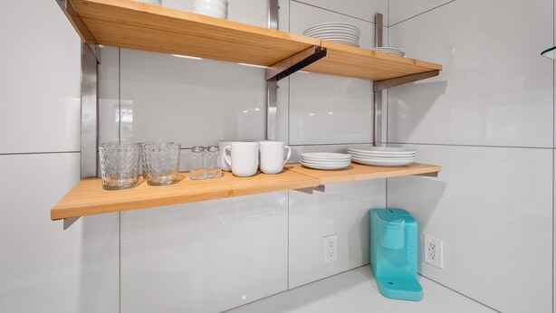 Une cuisine avec des armoires blanches, deux étagères en bois et une cafetière de couleur turquoise.