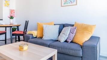 Una sala de estar moderna con un sofá gris, varios cojines, una mesa de centro de madera y una zona de comedor con una silla y una mesa.