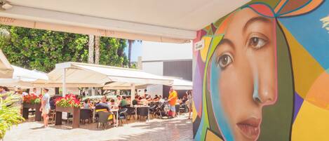 Un área de comedor al aire libre con mesas y sillas, rodeada por un mural.