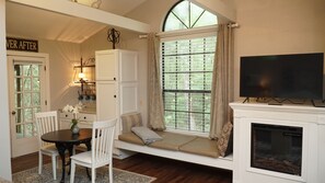 A cozy living room with a fireplace, a television, a dining area with a table and chairs, and a large window with blinds.