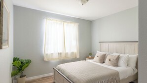 Une chambre à coucher avec un lit, une table de chevet, une plante en pot et une fenêtre ornée de rideaux légers.