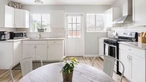 Une cuisine moderne avec des meubles blancs, une table à manger avec des chaises, une plante en pot et une cuisinière.